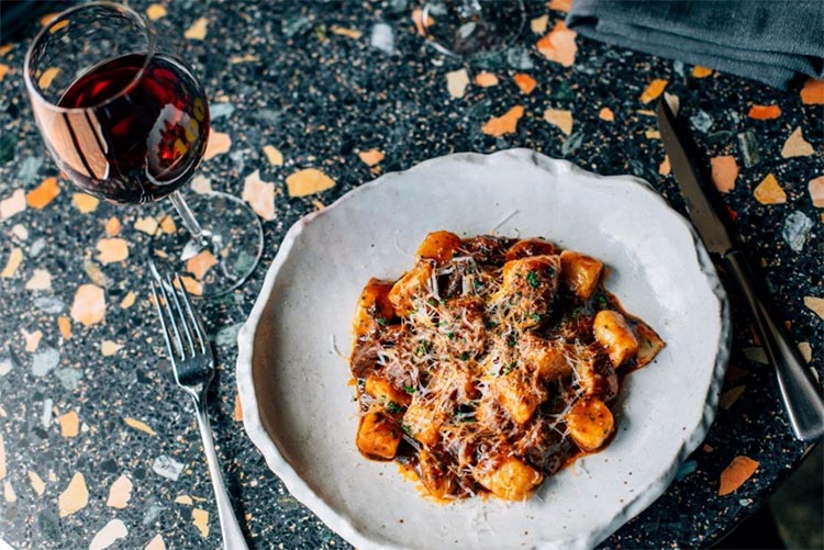 a bowl of gnocchi sitting on a table beside a glass of red wine